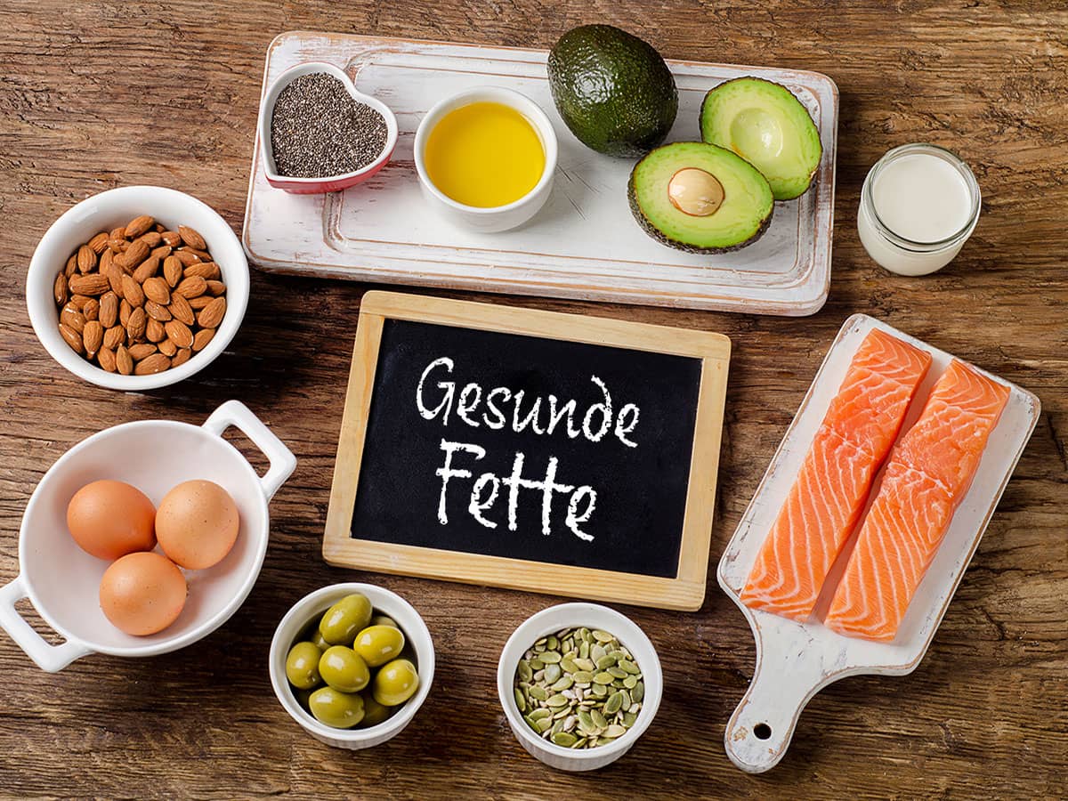 Gesunde Fette Eine kleine Tafel mit der Aufschrift 'Gesunde Fette'. Um die Tafel herum sind diverse Beispiele für Gesunde Fette, wie zum Beispiel Lachs, Avocado oder Eier.