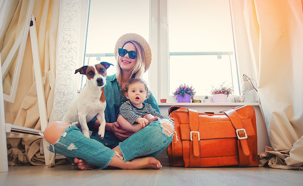 Reisevorbereitung mit Hund Eine Frau sitzt mit Hund und Kind im Arm, neben einer gepackten Reisetasche.
